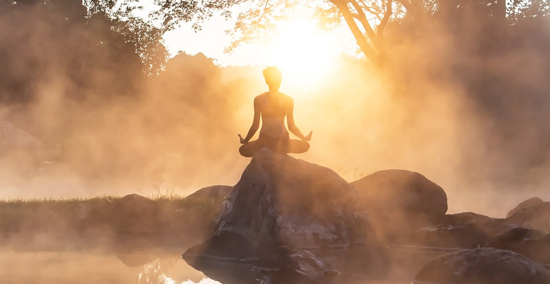 A person meditating on a rock near water on a misty morning. 