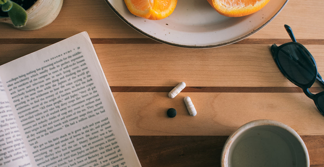 Two herbal supplements sitting next to each other on a wooden desk, surrounded by a book, plate of oranges, sunglasses, and cup. 